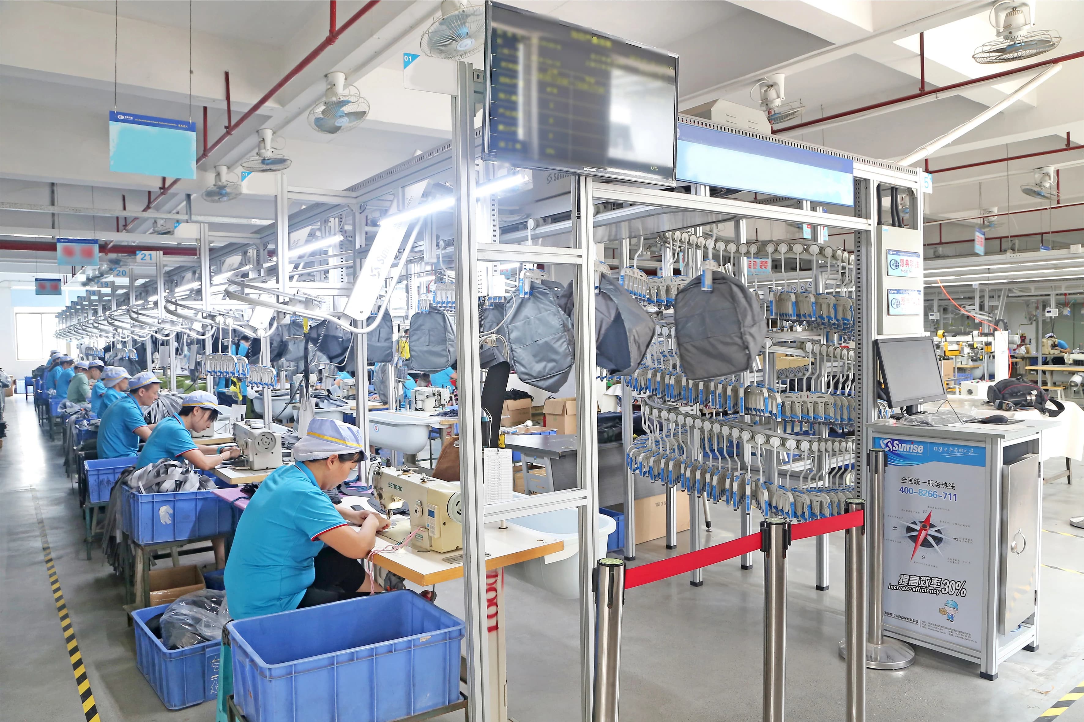 Team members sewing bags at workstations alongside an overhead conveyor on the production floor