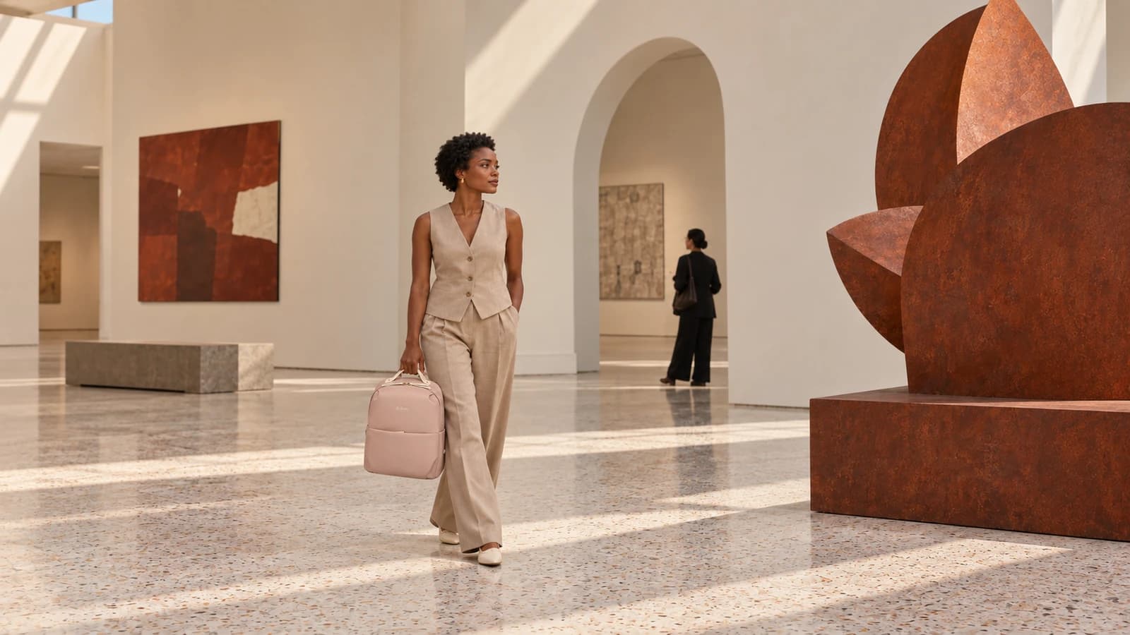 A woman in a tailored linen outfit carries a pink backpack by its top handle through an art gallery